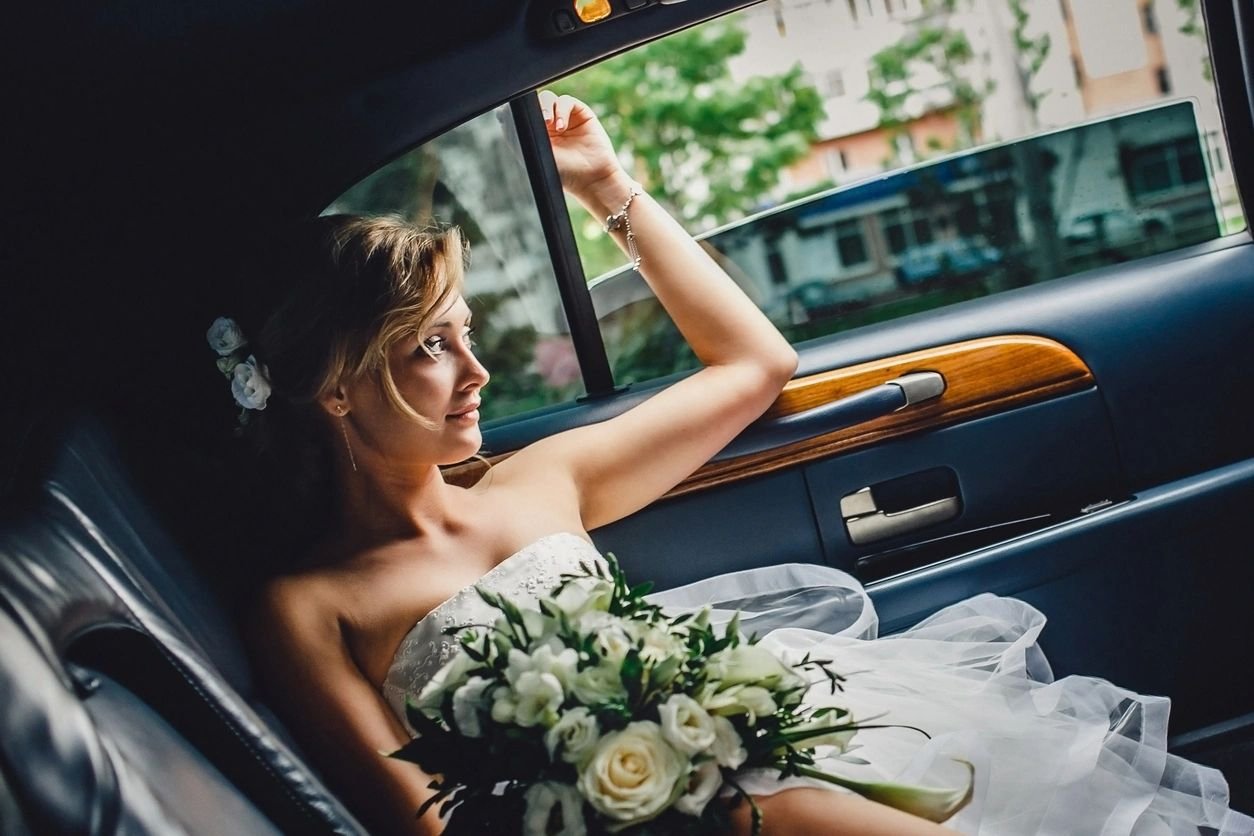 Portrait of a beautiful blonde bride with bouquet sitting in  wedding car and looking at somebody or something through the window. Wedding day. Daylight. (Portrait of a beautiful blonde bride with bouquet sitting in  wedding car and looking at somebod